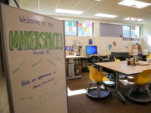 A peek in the Makerspace door at the whiteboard welcoming patrons to the space and a slightly blurred view of some of the machines available in the space such as the laser cutter and a large touch screen drawing computer