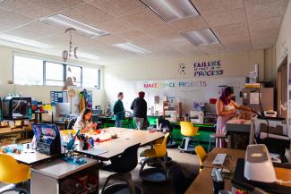 Wide shot of the Makerspace room - it's very full of stuff, including 3D printers, sewing machines, a button maker, and large worktables in the center. A students sits at the center table working on a project, behind her two people work at the embroidery machine, and on the right side of the room a student cuts fabric. There are signs on the back wall that say "Makerspace Projects" and "Failure is part of the process".